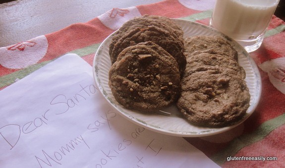 A holly-trimmed plate of homemade Flourless Pecan Sandies with a glass of milk and a note for Santa Claus placed on a Santa Claus dish towel on a white-washed table.