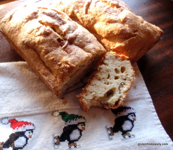 Two loaves of gluten-free Cinnamon Swirl Bread/Coffee Cake with end slices off of one, all sitting on wooden sideboard and cross-stitched towel with skating penguins.