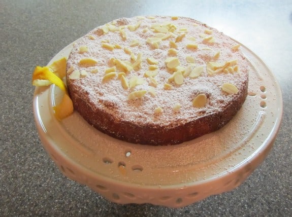 Flourless Gluten-Free Orange Almond Cake dusted with powdered sugar and topped with sliced almonds. Served on a white milk glass cake stand with a twist of orange zest. Recipe from Laura Uyemura, RDcipe 