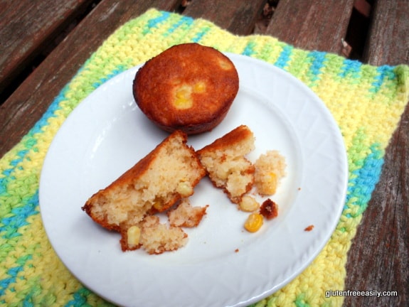 Gluten-Free Tender Corny Almond Muffins on a small white plate on colorful crocheted dish cloth on a rustic picnic table.