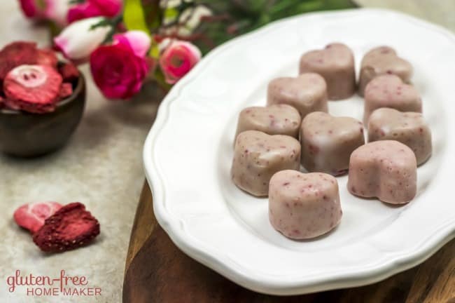Heart-shaped pale pink fudge candies on a white oval plate with a vase of flowers.