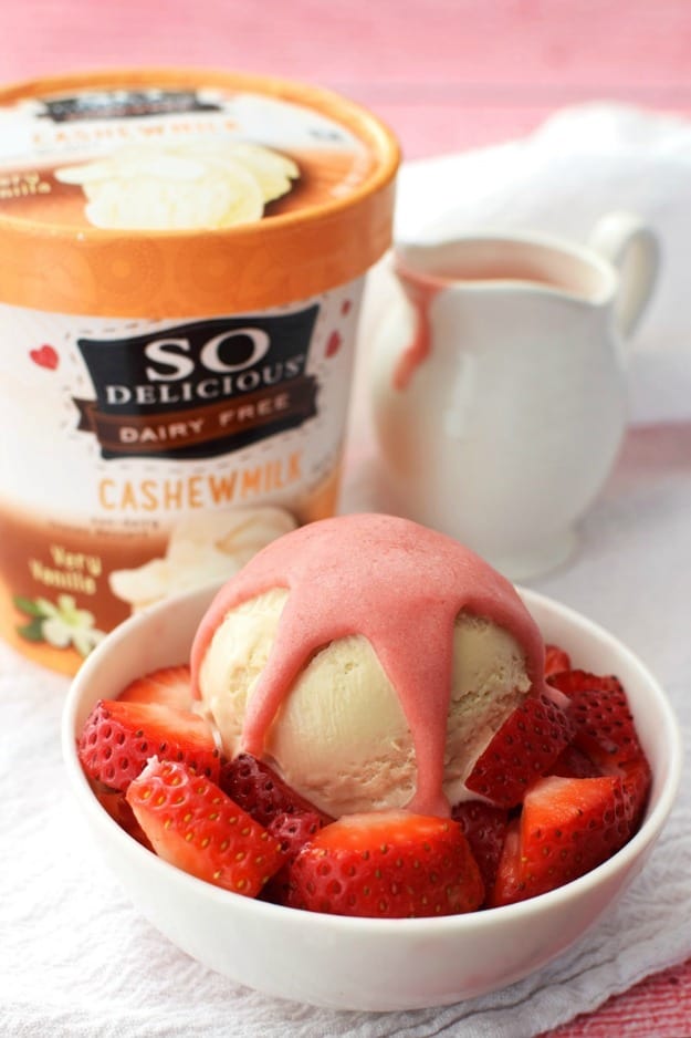 Pink strawberry topping on a scoop of vanilla ice cream surrounded by strawberry slices in a white bowl with a carton of ice cream beside it.