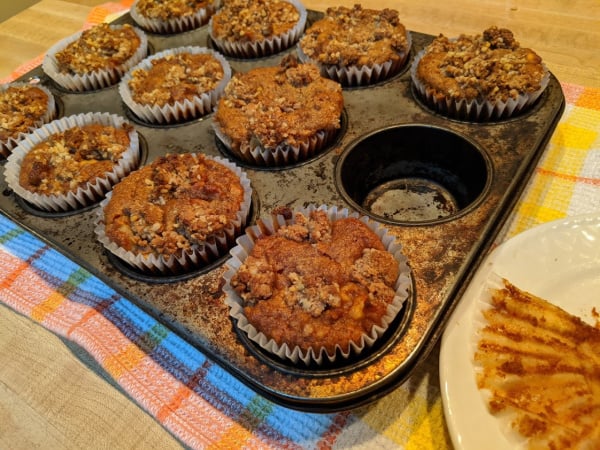 Gluten-free and paleo Apple Crumb Muffins in old Ecco muffin tin cooling on plaid cotton dish towel. One muffin already eaten.