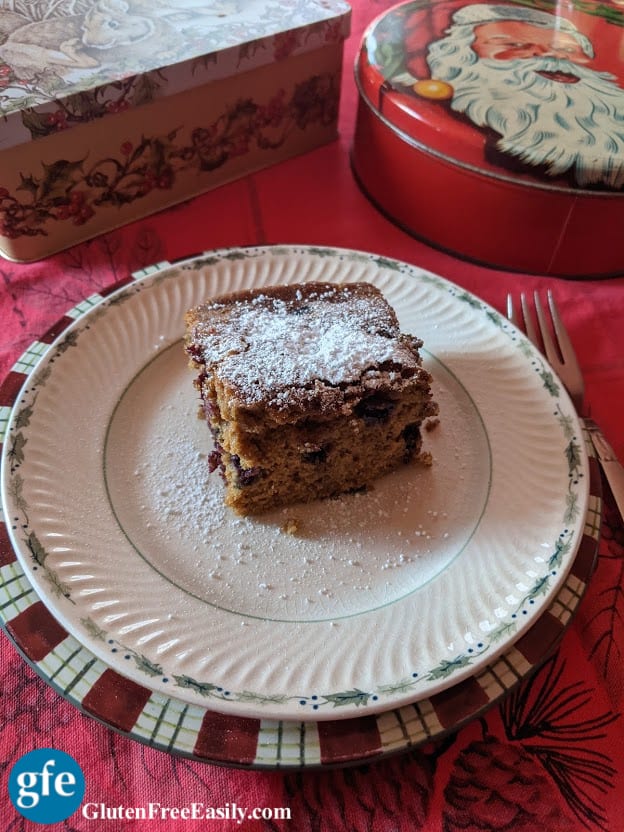 Single slice of Gluten-Free Blueberry Gingerbread with a dusting of powdered sugar. Served on two Christmas plates with fork and Christmas tins.