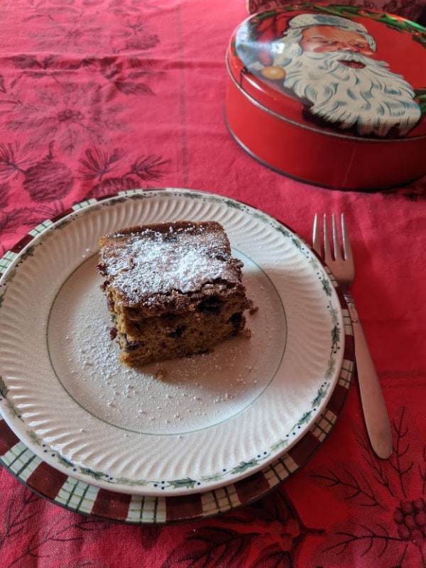 Single slice of Gluten-Free Blueberry Gingerbread with a dusting of powdered sugar. Served on two Christmas plates with fork and Christmas tins.