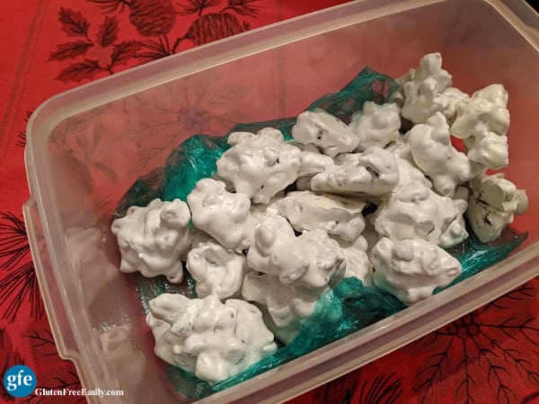 Naturally gluten-free Forgotten Cookies nestled in green plastic wrap in a Tupperware bread container sitting on a holiday tablecloth.