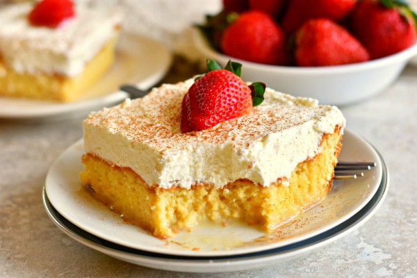 Gluten-Free Tres Leches Cake from Mama Knows Gluten Free. One slice on white plate with bite taken out of it and another slice behind it with a bowl of strawberries beside both.