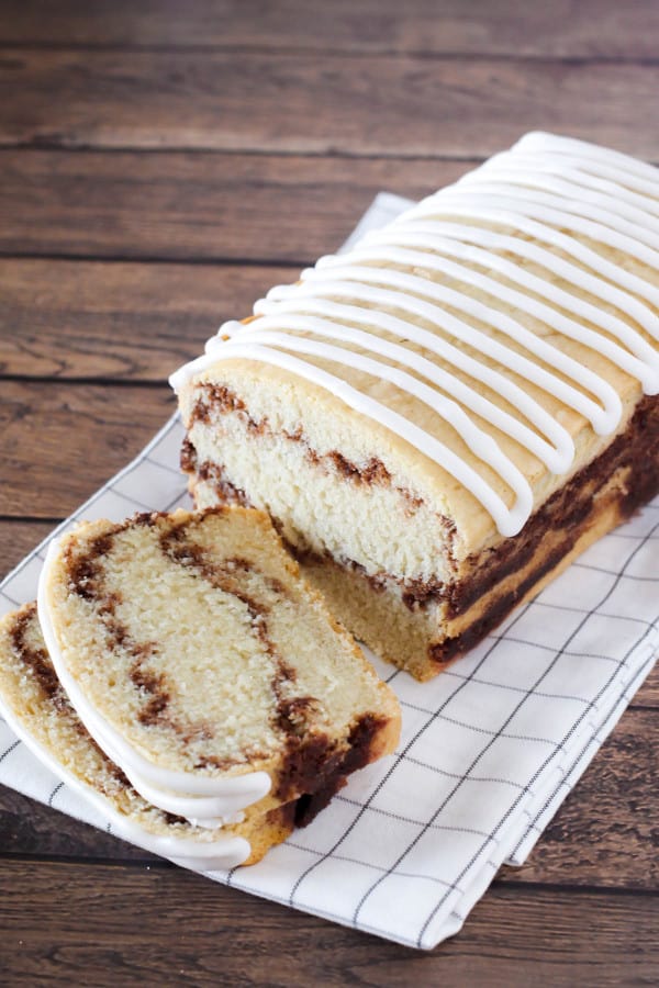 Loaf of gluten-free Cinnamon Swirl Bread with ribbon icing and two slices laying down on black and white plaid dish towel on hardwood surface.