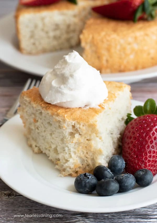 Gluten-Free Angel Food Cake. One of the best gluten-free angel food cake recipes featured on gfe. A wedge slice topped with a dollop on whipped cream on a white plate with blueberries and a strawberry. Behind it is the whole cake topped with berries with the wedge slice missing.