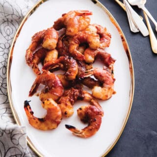 Gluten-Free Prosciutto-Wrapped Glazed Shrimp on oval white gold-rimmed platter on gray table with silver and table forks and white print scarf.