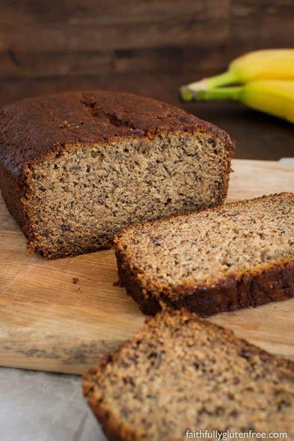 Gluten-Free Banana Bread from Faithfully Gluten Free. One of the best gluten-free banana bread recipes featured on gfe. Loaf with two slices cut on wooden cutting board with bunch of two bananas nearby.