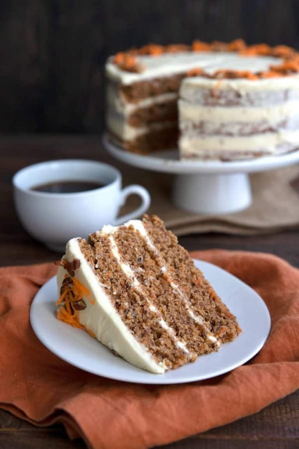 Gluten-Free Classic Keto Carrot Cake on cake stand with slice cut out on plate sitting on napkin and a cup of coffee. One of the featured gluten-free carrot cake recipes features on gfe.