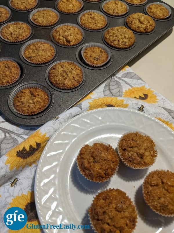 A muffin tin full of gluten-free Pecan Pie Mini Muffins with four more mini muffins on a white plate on a sunflower dish towel.