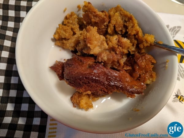 A serving of Gluten-Free Pumpkin Bread Pudding topped with almond milk in a white bowl on a black gingham dish towel.