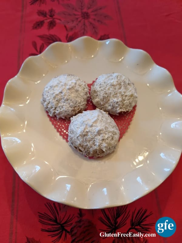 Close-up of Gluten-Free Swedish Pecan Ball Cookies on Ruffled White Dessert Stand on Vintage Red Holiday Tablecloth with holly and poinsettias.