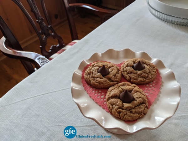 Gluten-Free Flourless Peanut Butter Blossom Cookies on elevated white ruffled dessert plate on dining room table with end chair showing.