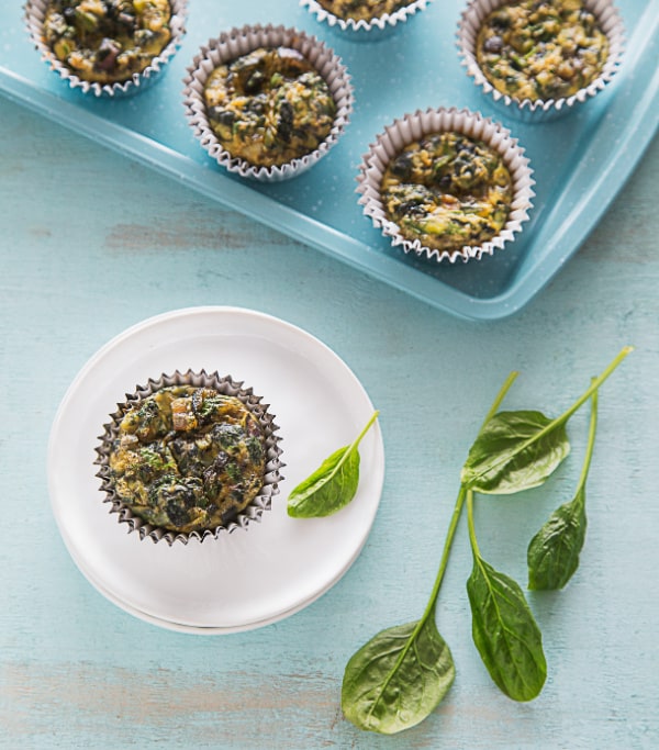 Gluten-Free Egg Muffins with Spinach (Flourless) from Dr. Becky Campbell. Single muffin on white sauce with spinach leave beside it on plate and counter with rest of muffins cooling in muffin tin.