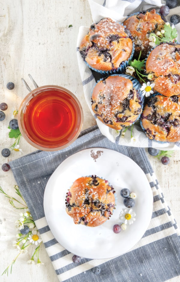 Gluten-Free Blueberry Muffins with one on a white plate on a dish towel, others on a whlte plate, and all beside a cup of tea. From TheMigraine Relief Plan (MRP) Cookbook. MRP compliant. Migraine friendly.