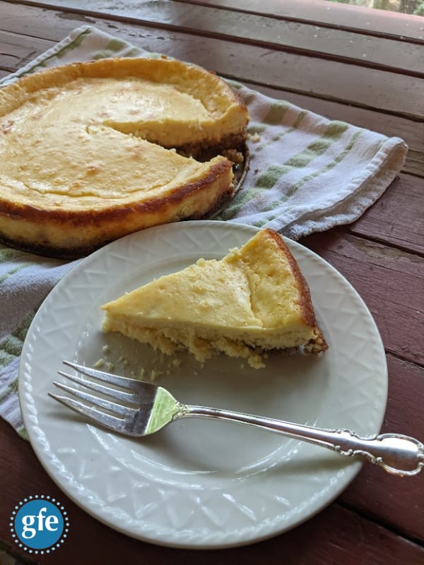 Gluten-Free Creamy Vanilla Cheesecake on green and white striped towel on picnic table with a single slice on a small white plate with silver dessert fork.