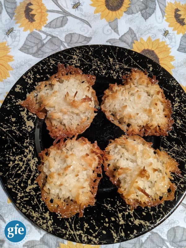 Flourless Gluten-Free Coconut Macaroons made with Bakers Angel Flake Sweetened Coconut. Four cookies on a black plate with gold splashes on a sunflower dish towel.