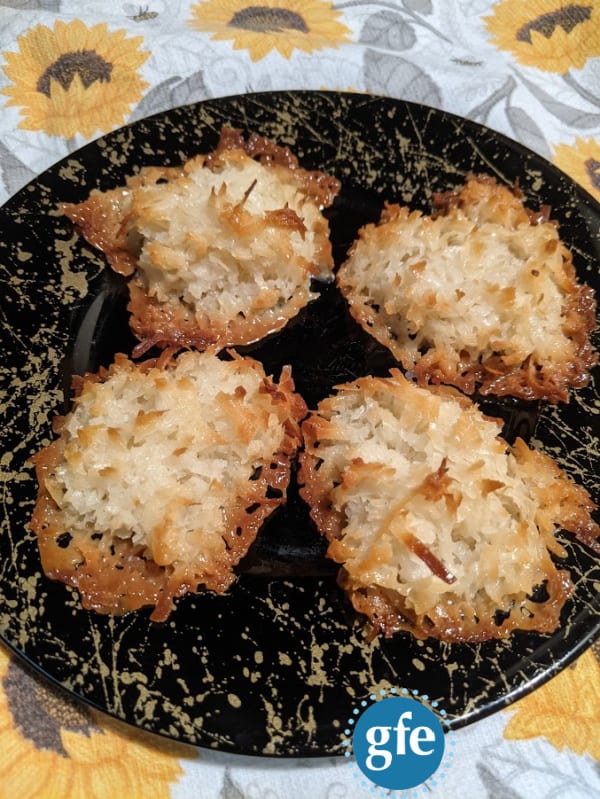 Flourless Gluten-Free Coconut Macaroons made with Bakers Angel Flake Sweetened Coconut. Four cookies on a black plate with gold splashes on a sunflower dish towel.