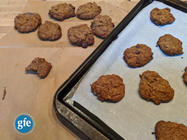 Flourless Gluten-Free Cashew Butter Coconut Chocolate Chip Cookies. Some cookies including a half eaten on cooling on brown paper and more still setting up on parchment-lined baking sheet.
