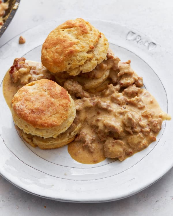 Gluten-Free Biscuits and Sausage Gravy from Mama Knows Gluten Free on a shiny white plaqte rimmed with silver beside a skillet of sausage gravy.