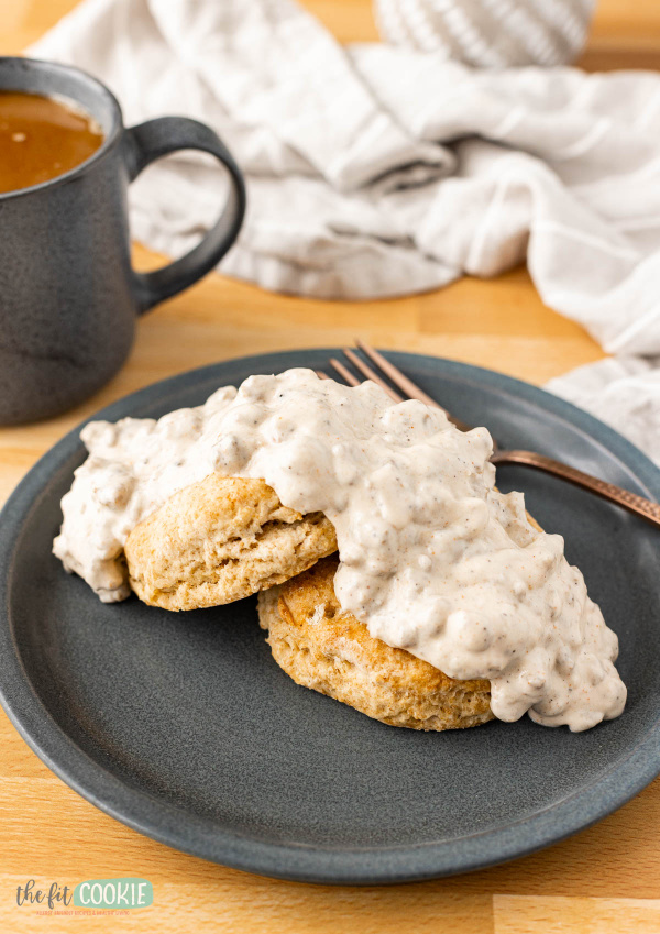 Gluten-Free Biscuits and Sausage Gravy from The Fit Cookie on a green plate with a fork and a green mug of coffee and a dish towel nearby.