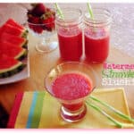 Watermelon-Strawberry Slushies in glasses beside a plate of watermelon slices and a bowl of strawberries on a colorful striped placemat.