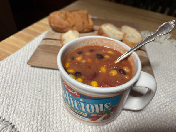 Quick and Easy Four-Bean Vegetarian Chili in a vintage Campbell's soup mug with an ornage silver spoon on an off white placemat beside a cutting board with a gluten-free baguette.