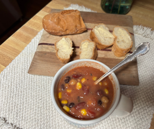 Quick and Easy Four-Bean Vegetarian Chili in a vintage Campbell's soup mug with an ornage silver spoon on an off white placemat beside a cutting board with a gluten-free baguette.