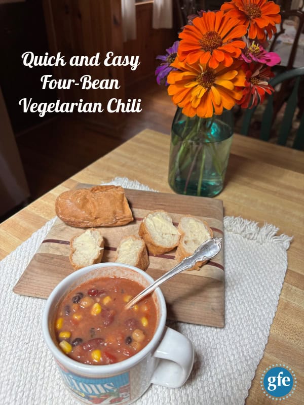 Quick and Easy Four-Bean Vegetarian Chili in a vintage Campbell's soup mug with an ornage silver spoon on an off white placemat beside a cutting board with a gluten-free baguette and cut zinnias in a blue Ball jar.