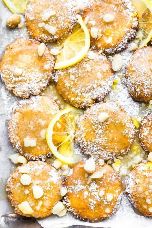Gluten-Free Lemon Glazed Macadamia Nut Cookies from Cotter Crunch on a parchment-lined baking sheet with a sprinkle of powdered sugar, macadamia nuts, white chocolate chips, and lemon slices.