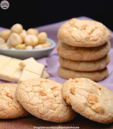 Gluten-Free White Chocolate Macadamia Cookies stacked domino effect from The Gluten and Dairy Free Bakehouse on a table with a pink napkin to the side topped by a stack of cookies, white chocolate squares, and a saucer of macadamias.