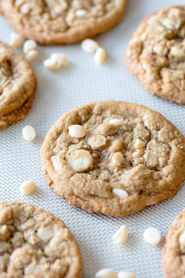 Gluten-Free White Chocolate Macadamia Nut Cookies from Allergy Free Alaska spread on a white textured tablecloth with white chocolate chips scattered about.